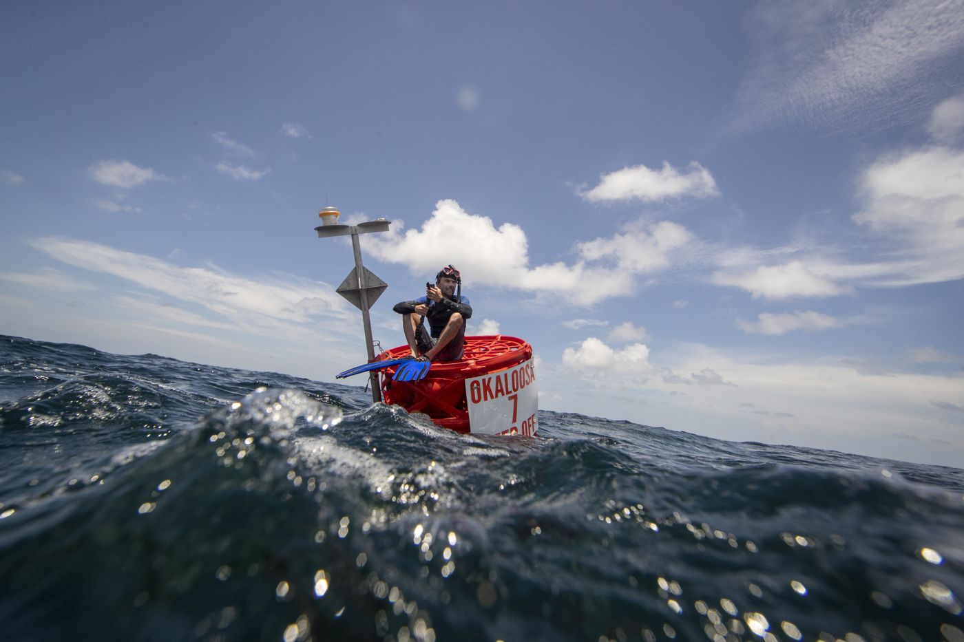 A man sitting on a red buoy offshore under a blue sky