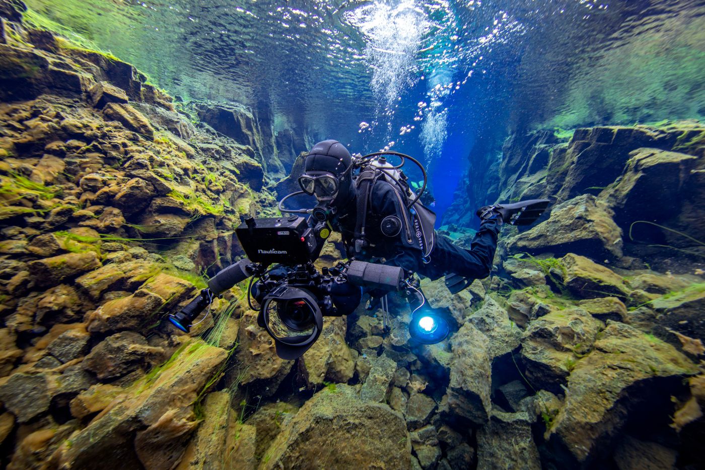A diver filming with an underwater camera rig between rock walls in clear blue water
